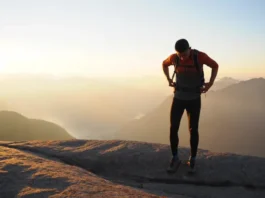 A hiker stands on a mountain summit at sunset, thoughtfully adjusting their large backpacking backpack before continuing their journey.