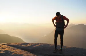 Choosing a Backpacking Backpack: A Hiker’s Skill Guide A hiker stands on a mountain summit at sunset, thoughtfully adjusting their large backpacking backpack before continuing their journey.
