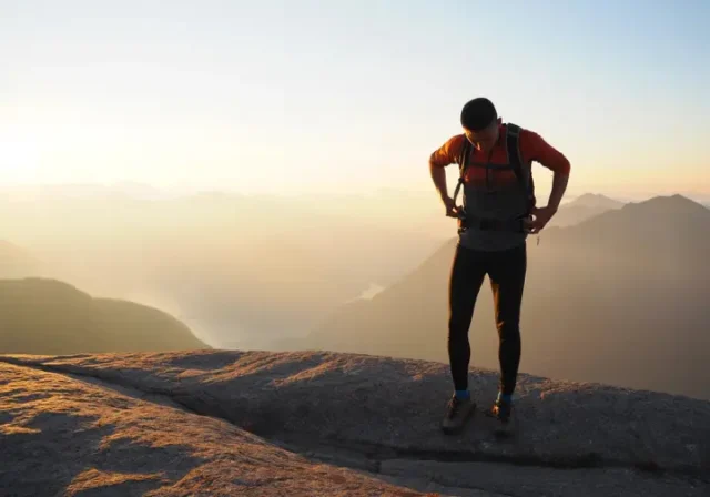 A hiker stands on a mountain summit at sunset, thoughtfully adjusting their large backpacking backpack before continuing their journey.