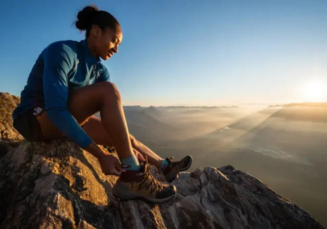 A female hiker adjusts her hiking boot and technical boot sock while pausing on a mountain overlook with a stunning view at sunrise.