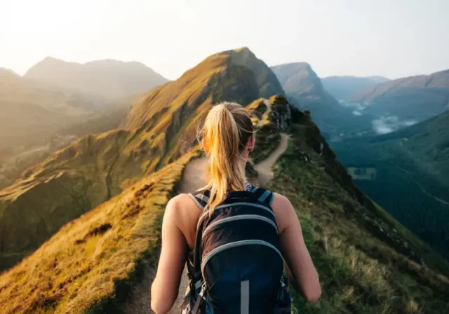 A hiker wearing a daypack stands at a trail junction in the mountains, choosing between a sunny ridge and a misty valley, symbolizing the selection of the right rucksack for any trail.