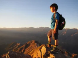 A young child in hiking boots stands on a mountain summit at sunset, looking out over a beautiful valley, embodying the spirit of adventure.