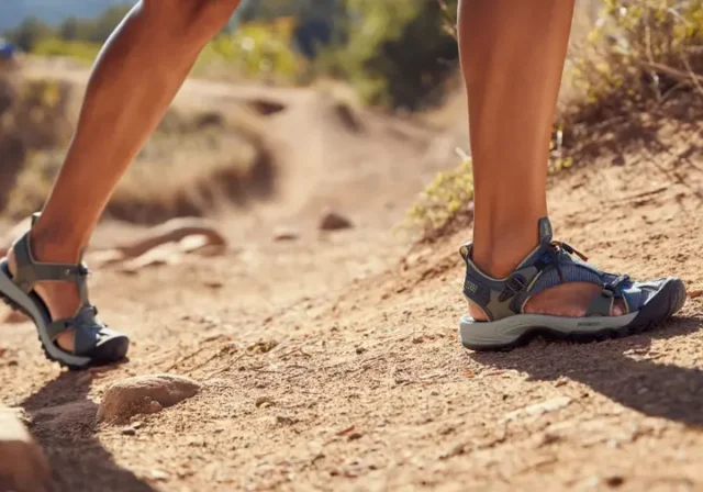 A female hiker compares a zero-drop hiking sandal on one foot with a high-drop hiking sandal on the other, illustrating the choice between footwear philosophies for women.
