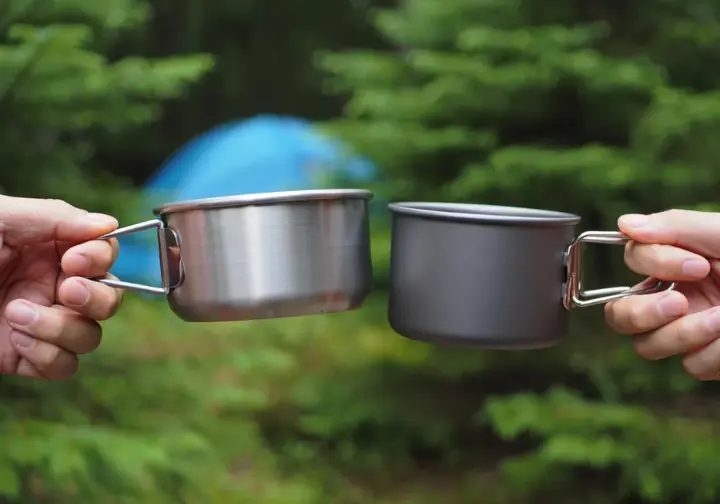 A close-up of a hiker's hands comparing an ultralight titanium camping pot to a heavier hard-anodized aluminum pot at a campsite.