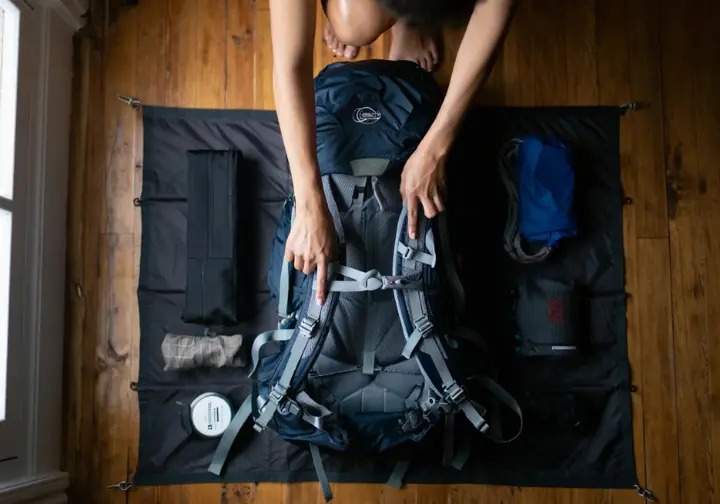 A top-down view of hiking gear laid out neatly, with a hiker's hand pointing to the crucial suspension system on a modern backpacking backpack.