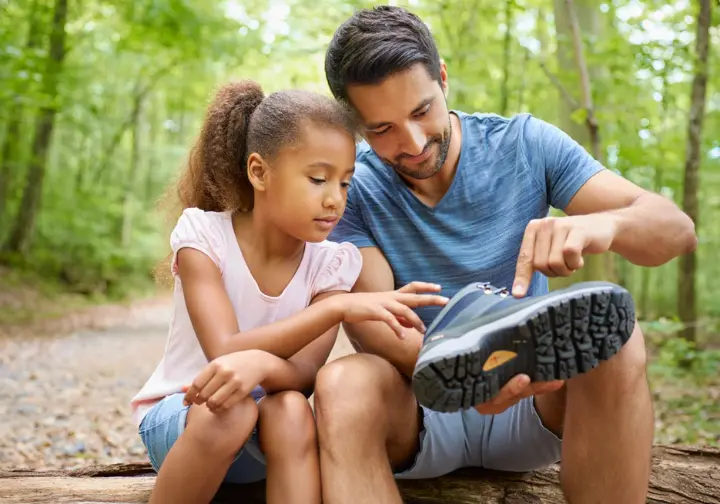 A father sits with his young daughter on a trail, showing her the traction on the bottom of her new hiking boot.