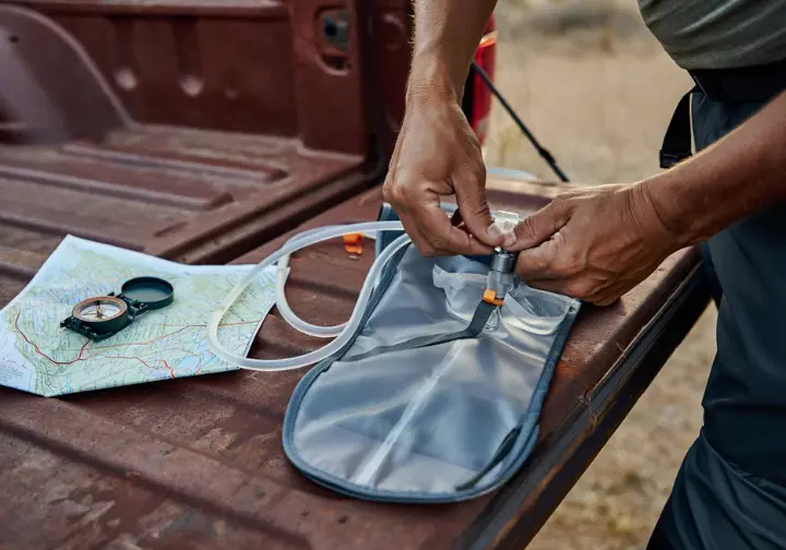 A hiker's hands carefully inspect the fundamental features of a hydration pack bladder laid out with a map and compass before a hike.