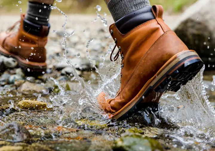 A close-up, low-angle action shot of a hiker's boots splashing through a stream, demonstrating the real-world testing of boot socks.