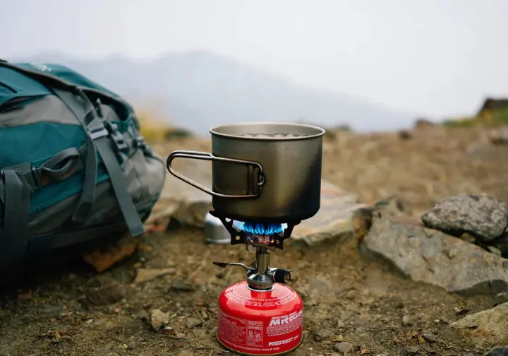 An MSR PocketRocket 2 camp stove boiling water in a pot during a field test on a rocky, misty mountain trail.