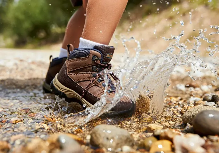 A close-up action shot of a child's muddy hiking boot splashing through a creek, demonstrating field testing.
