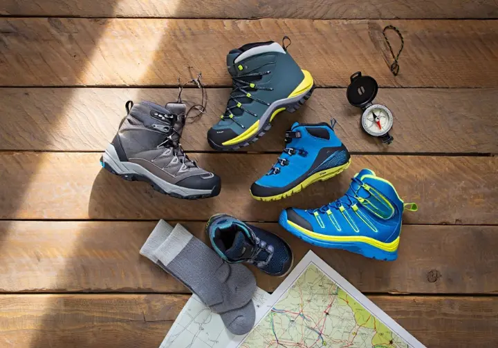A flat lay of three different pairs of the best kids' hiking boots for 2025, arranged on a wooden background with a map and socks.