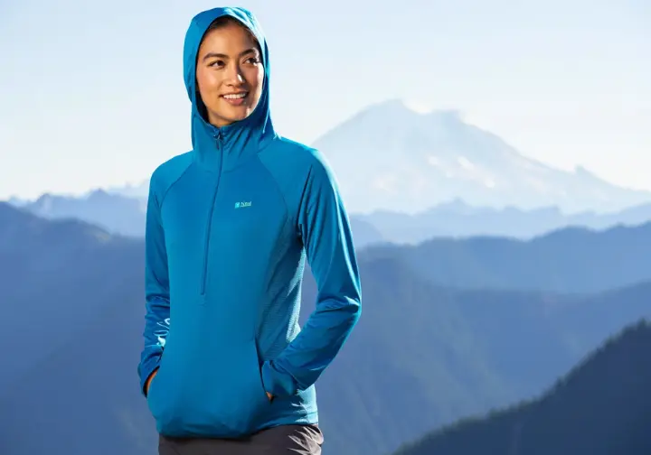 A confident female hiker wearing a high-performance sun hoodie smiles at a scenic mountain viewpoint, representing a top pick for best women's hiking shirts.