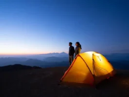 A glowing 2-person backpacking tent sits on a mountain overlook at dusk, as two hikers admire the vast, layered mountain vista, representing the adventure that lies beyond the gear specs.