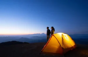 Beyond the Specs: A Guide to 2 Person Backpacking Tents A glowing 2-person backpacking tent sits on a mountain overlook at dusk, as two hikers admire the vast, layered mountain vista, representing the adventure that lies beyond the gear specs.