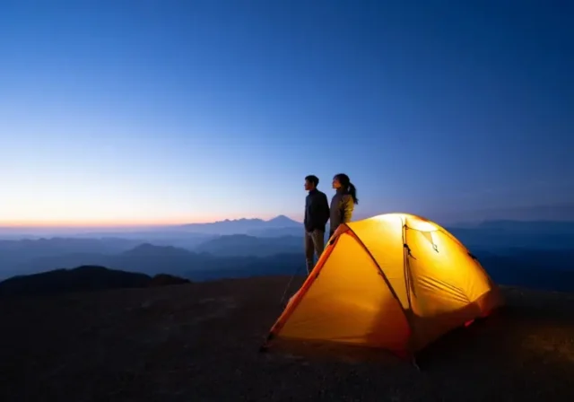 A glowing 2-person backpacking tent sits on a mountain overlook at dusk, as two hikers admire the vast, layered mountain vista, representing the adventure that lies beyond the gear specs.