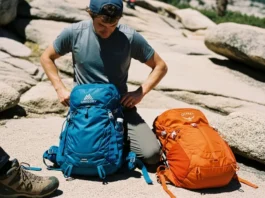 A hiker pauses on a sunny, rocky trail to adjust the hipbelt of their Gregory backpack, with an Osprey pack resting on the ground nearby.