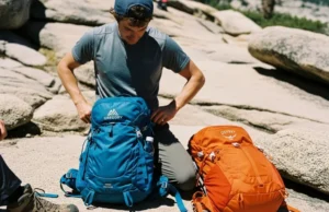 Gregory vs Osprey 2025: The Hiker’s Load-Comfort Verdict A hiker pauses on a sunny, rocky trail to adjust the hipbelt of their Gregory backpack, with an Osprey pack resting on the ground nearby.