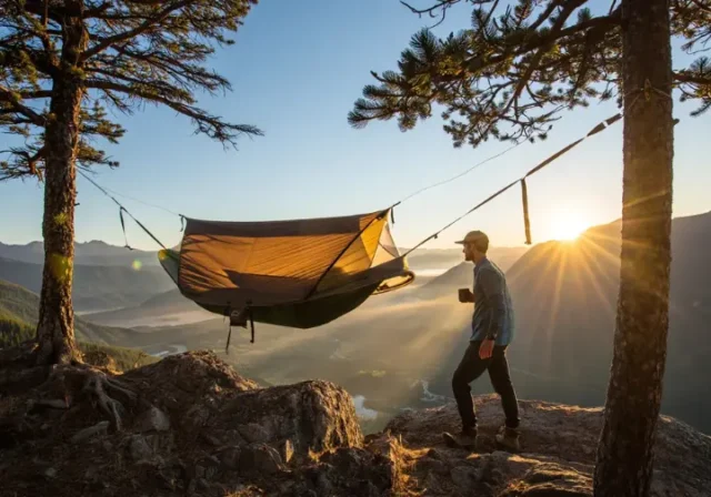 A hiker enjoys a morning coffee next to their field-tested hammock tent, set up on a mountain overlook at sunrise.