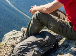 A hiker wearing dusty outdoor pants sits on a rock overlooking a mountain valley in the harsh midday sun.