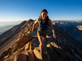 A female hiker wearing performance hiking shorts strides confidently along a dramatic, sunlit mountain ridge, embodying a trail-rated gear review.