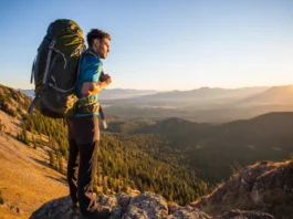 A hiker with a perfectly fitted backpack stands on a mountain summit at sunset, looking out over a vast valley.