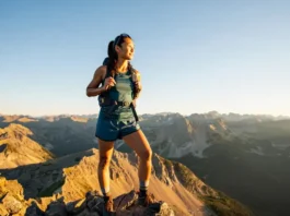 A female hiker wearing functional hiking shorts stands on a mountain summit at golden hour, looking out over a vast mountain range.