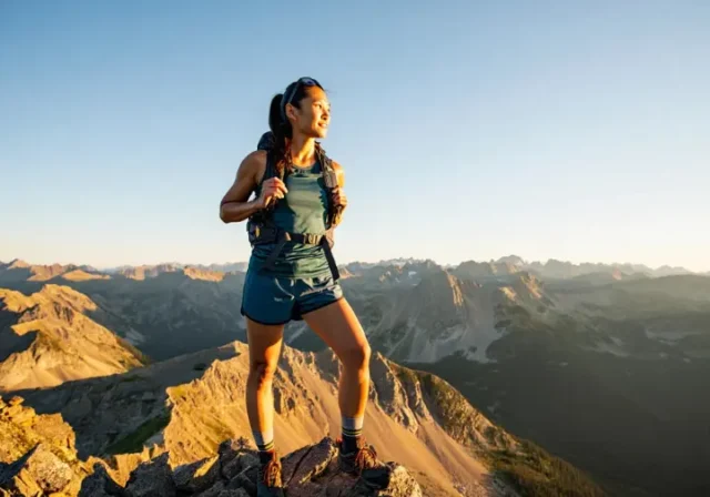 A female hiker wearing functional hiking shorts stands on a mountain summit at golden hour, looking out over a vast mountain range.