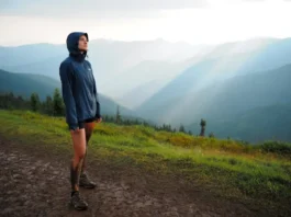 A female hiker wearing waterproof hiking shoes stands on a muddy mountain trail, looking out at a scenic, misty landscape.