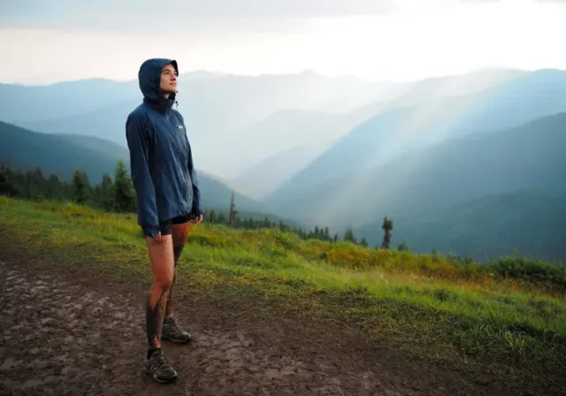 A female hiker wearing waterproof hiking shoes stands on a muddy mountain trail, looking out at a scenic, misty landscape.