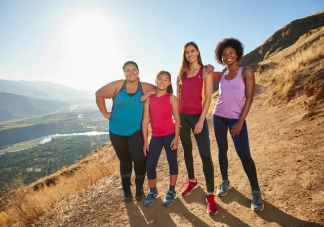 A diverse group of four women hikers with different body types standing confidently on a mountain summit, all wearing well-fitting hiking pants.