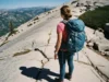 Women’s Water-Resistant Boots 2025: Fit & Trail Matrix Guide A woman hiker in water-resistant boots and a backpack pauses on a sunny, rocky trail, seen from a wide, side-angle view.