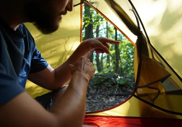 A hiker's hands point out a construction detail inside a 2-person backpacking tent, with the open door framing a sunny forest view, illustrating the process of evaluating what truly matters in a tent.