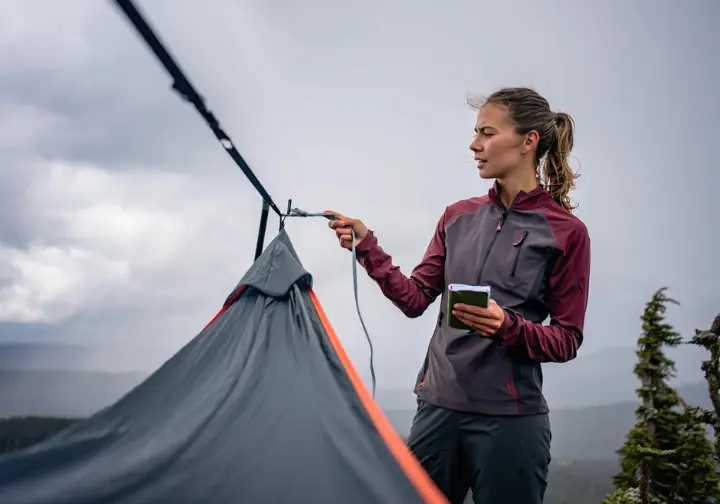 A field-tester makes adjustments to a hammock tent on a windy, overcast ridgeline, demonstrating the rigorous testing process.