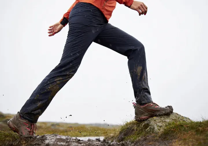 A female hiker field-testing hiking pants by high-stepping over a muddy rock on a rainy, challenging trail, demonstrating mobility and durability.