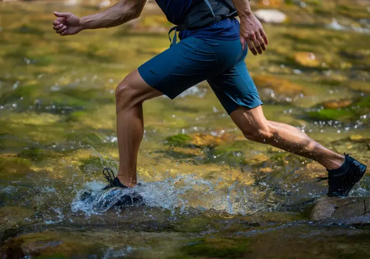 A hiker tests the water resistance and mobility of his hiking shorts while crossing a rocky stream, demonstrating the real-world testing protocol.