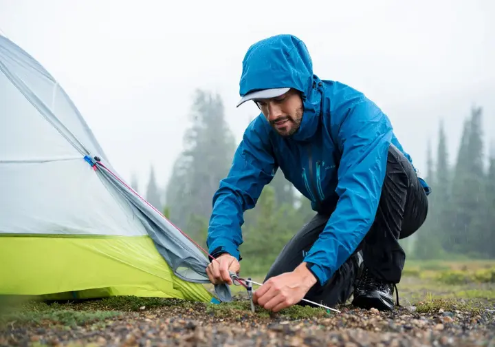 A hiker in a rain jacket carefully tests a 2-person backpacking tent by setting it up on rocky, wet ground in a misty forest, demonstrating a rigorous real-world review process.