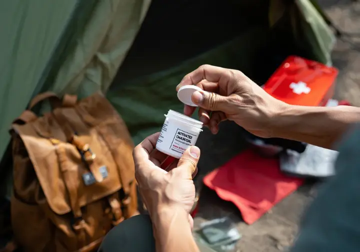 Hands opening activated charcoal from a first-aid kit at a campsite, showing its emergency use for specific situations in activated charcoal camping.