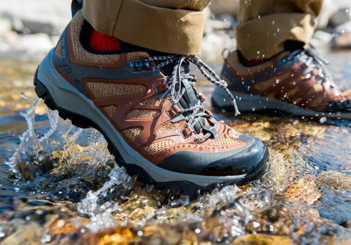 A close-up action shot of a woman's waterproof hiking shoe splashing into a stream, demonstrating its performance during testing.