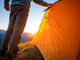 A solo backpacker stands in full hiking gear next to their pitched tent in a mountain meadow during a stunning sunset