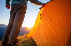 Backpacking with Tent: 2025 Field-Tested Guide to Pack & Pitch A solo backpacker stands in full hiking gear next to their pitched tent in a mountain meadow during a stunning sunset