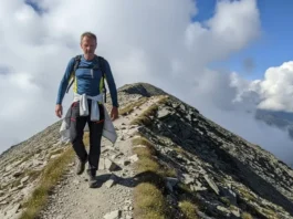 A full-body shot of a man hiking on a sunny, windswept mountain trail, wearing technical hiking trousers.