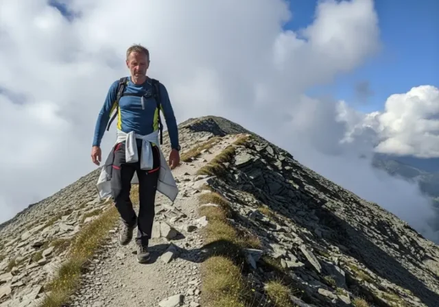 A full-body shot of a man hiking on a sunny, windswept mountain trail, wearing technical hiking trousers.