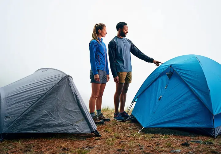 Two hikers stand in full gear comparing a freestanding tent and a non-freestanding tent pitched next to each other in a field.
