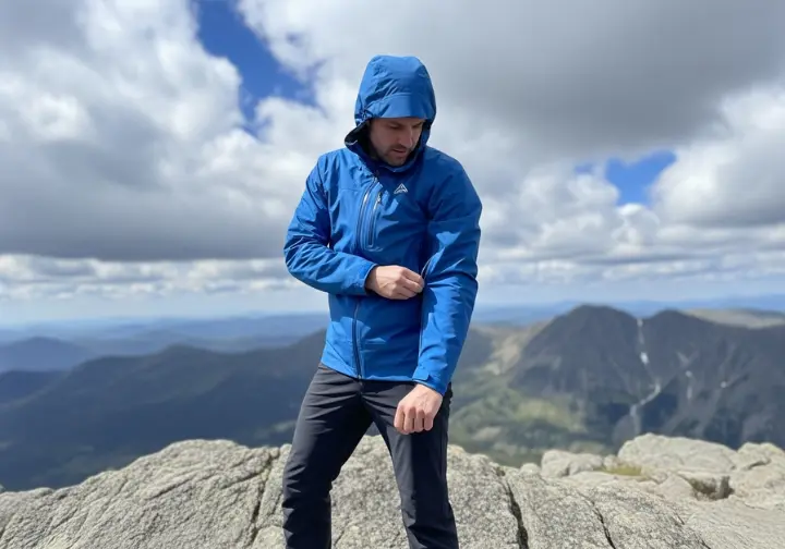 A full-body shot of a hiker standing on a rock, inspecting the pit-zip vent on his blue waterproof rain jacket.
