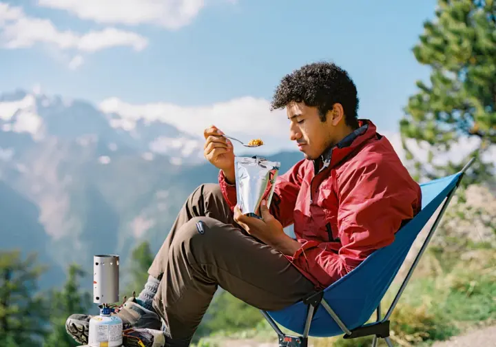 A hiker enjoys an easy, rehydrated meal from a pouch at a scenic mountain campsite, demonstrating lightweight meal strategies from a hiker's cookbook.