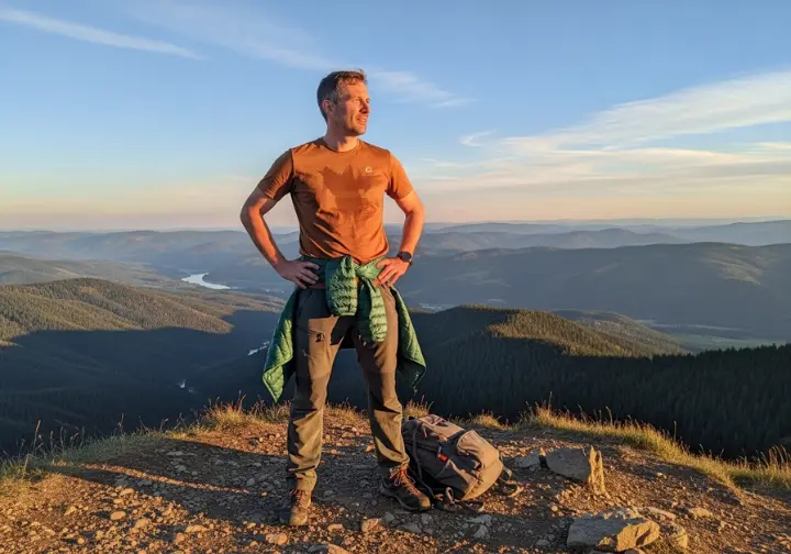 A full-body view of a hiker in technical trousers standing on a mountain peak at sunset, looking out over a vast valley.