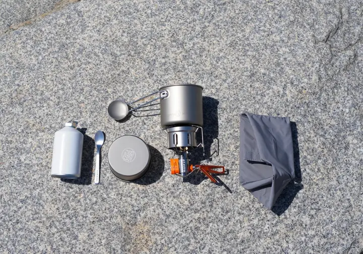 Flat lay of ultralight trail kitchen gear including a compact stove, titanium pot, and long spoon, showing essential equipment for easy camping meals.