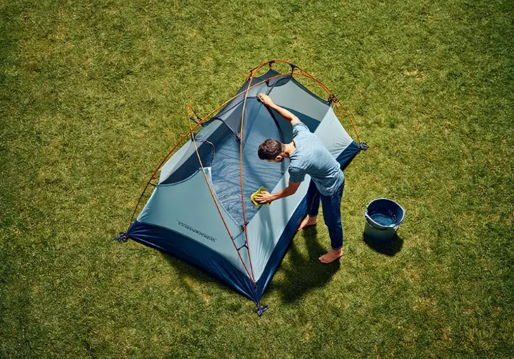 A person in their backyard carefully cleans their backpacking tent, which is pitched on the grass to air dry before storage.
