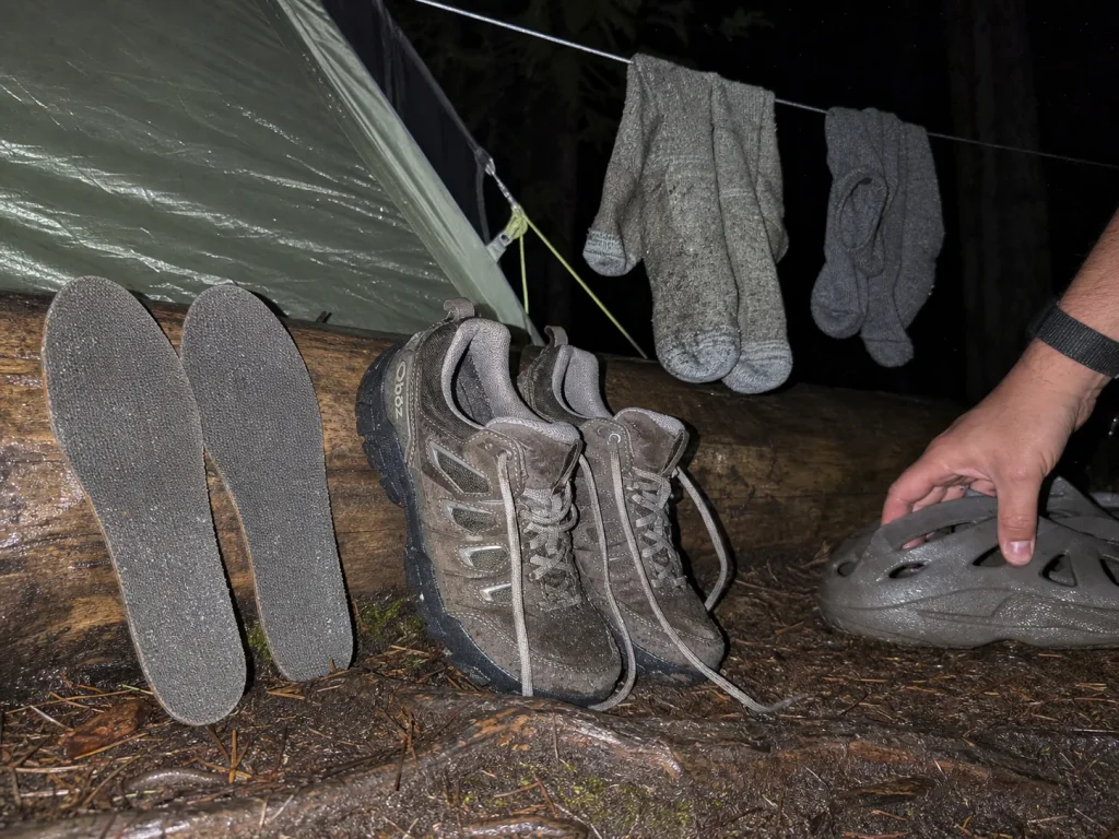 Wet backpacking shoes propped open to dry at camp at dusk beside dry camp shoes