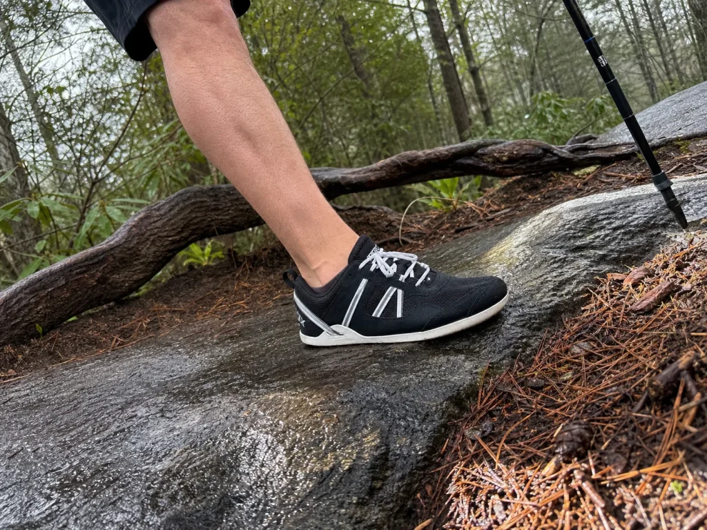 Hiker's foot in a minimalist sneaker placed on off-camber wet rock on a trail